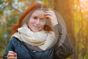 Portrait of a red-haired smiling girl in a jacket and scarf straightening her hair against the background of autumn nature and the