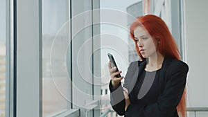 Portrait of a red-haired woman holding a mobile phone in her hand looking gloomily out the window. Angry woman in the
