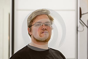 Portrait of a red-haired young man with glasses and a beard on a light neutral background in the room.