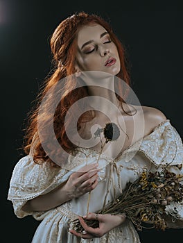 Portrait of a red-haired young woman in a vintage gold dress with a dry bouquet in hands on a black background. A princess. Fairy