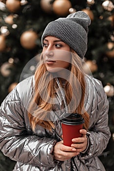 Portrait of a red-haired young woman in a vintage knitted gray hat in a fashionable silver jacket with a cup of coffee in hands