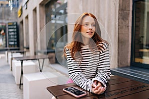 Portrait of redhead young woman relaxing in cafe at table on city street, looking away. Happy caucasian woman in casual