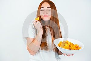 Portrait of redheaded young woman biting crispy potatoe chips with pleasure looks happy standing on isolated white backgroung,