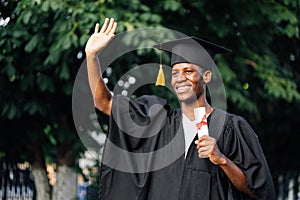 Portrait of rejoicing black guy graduate from university stand outdoors near trees waving hand and holding higher