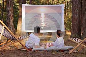 Portrait of relaxed loving couple lying in the forest with overhead projector, watching romantic movie, enjoying sunset on screen
