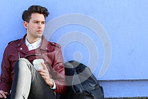 Portrait of a relaxed modern man sitting outdoors with cup of coffee and his backpack