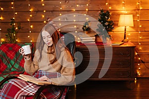 Portrait of relaxed woman holding cup with hot coffee and reading book while sitting at armchair