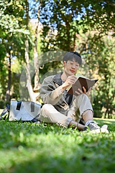 Portrait, Relaxed young Asian male college student chilling in the greenery park, reading a book