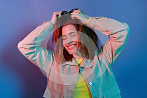 Portrait of relaxing happy young girl wearing casual jacket standing isolated over blue neon light background, dancing , raised