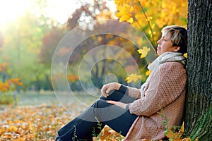 Portrait of relaxing woman in autumn city park. Sitting near tree. Bright sunlight and golden trees, fall season, yellow leaves