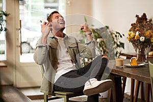 Portrait of relieved, happy young man, listens to happy news or podcast in earphones, sits in coffee shop, holds