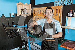 Portrait of a repairman in an apron leaning on a bicycle frame while holding a tablet