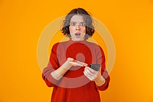 Portrait of resentful woman holding and using cell phone while standing isolated over yellow background