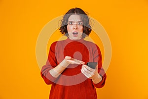 Portrait of resentful woman holding and using cell phone while standing isolated over yellow background photo
