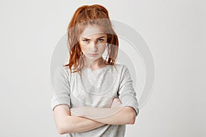 Portrait of resentful young pretty redhead girl looking at camera brutally with crossed arms over white background.