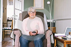 Portrait of retired elderly woman in comfortable armchair with TV remote control and watching television channel in