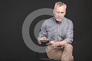 Portrait of retired senior man holding in his hand a digital tablet while sitting, holding glasses.  against black