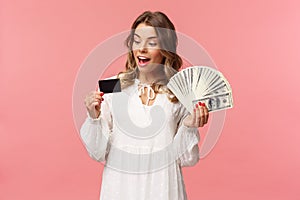 Portrait of rich good-looking blond girl in white dress, holding dollars money and credit card, winning lottery, think