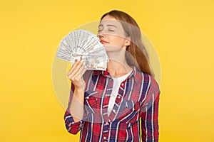 Portrait of rich greedy woman in casual checkered shirt holding money, smelling dollar banknotes with pleased expression