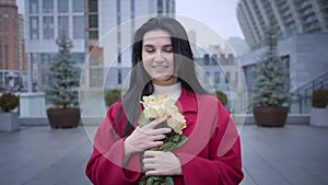 Portrait of romantic young Caucasian woman smelling yellow roses and looking at camera. Fashionable lady with black hair