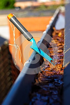 A portrait of a roof gutter full of fallen autumn leaves on a sunny day with a small blue garden shovel in it of someone who is