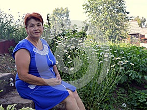 Portrait of 60s mature woman in blue dress sitting next to flower beds with daisies in village