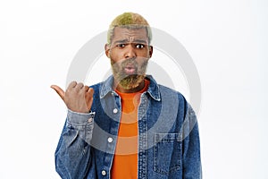 Portrait of sad african american guy, points left, shows smth disappointing and shocking, stands over white background