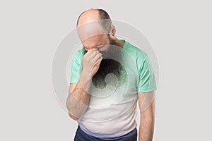 Portrait of sad alone depressed middle aged bald man with long beard in light green t-shirt standing holding his head down and