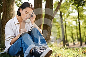 Portrait of sad asian girl writing in her diary and feeling uneasy, sitting in park alone under tree, expressing her
