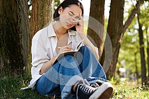 Portrait of sad asian girl writing in her diary and feeling uneasy, sitting in park alone under tree, expressing her