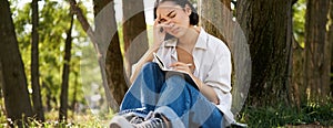 Portrait of sad asian girl writing in her diary and feeling uneasy, sitting in park alone under tree, expressing her