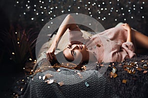 Portrait of sad attractive young woman with tinsel confetti and garland lights celebrating alonein dark room. New year`s