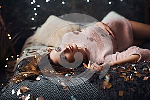 Portrait of sad attractive young woman with tinsel confetti and garland lights celebrating alonein dark room. New year`s