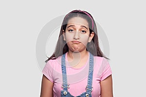 Portrait of sad beautiful brunette young girl in casual style, pink t-shirt and blue denim overalls standing with sadness and