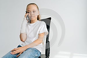 Portrait of sad blonde little girl with broken arm wrapped in plaster bandage talking smartphone, smiling looking down