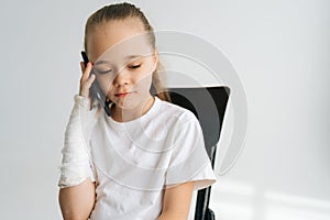 Portrait of sad blonde little girl with broken arm wrapped in plaster bandage talking smartphone, smiling looking down