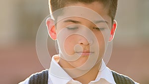 Portrait sad boy little child lonely pupil schoolboy face upset kid looks down with sadness stands outdoors on street