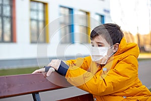 Portrait of a sad boy in a yellow jacket in a white medical mask sitting on a bench on the street