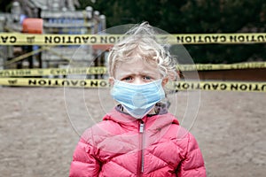 Portrait of sad Caucasian girl in face mask on closed playground outdoor. Kids play area locked with yellow caution tape in