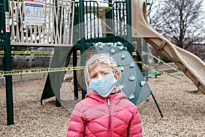 Portrait of sad Caucasian girl in face mask on closed playground outdoor. Kids play area locked with yellow caution tape in