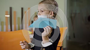 Portrait of sad Caucasian schoolgirl in Covid-19 face mask launching paper plane in classroom. Little Caucasian