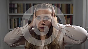 Portrait of sad caucasian woman who closes ears by hands sitting in room on bookshelf background