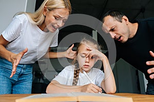 Portrait of sad cute little girl writing homework sitting at table on background of angry parents yelling and scolding