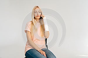 Portrait of sad depressed young woman with broken arm wrapped in plaster bandage talking mobile phone sitting on chair