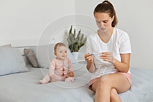 Portrait of sad frustrated young adult pregnant woman with pregnancy test posing in her bedroom with toddler baby, wearing white t