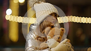 Portrait of sad girl lost in urban city at Christmas night. Stressed Caucasian kid hugging teddy bear looking around