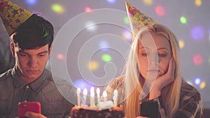 Portrait sad girl and a man in birthday hats sitting in front of cake with candles texting on their cellphones