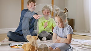 Portrait of sad little Caucasian girl sitting in living room with blurred boys laughing and pointing at background
