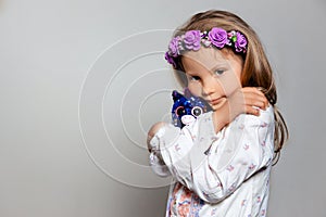 Portrait of sad little girl in white dress and purple wreath with plush unicorn toy on gray isolated background.