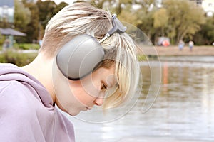 Portrait of sad lonely teenager boy sitting on the lake shore and listening to music on wireless headphones